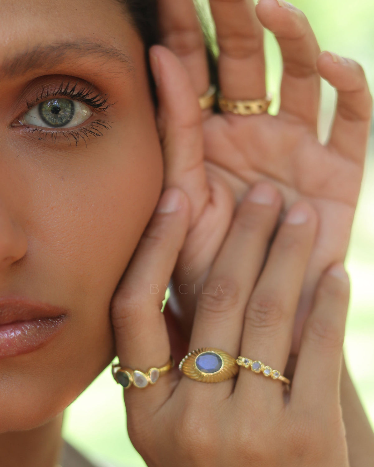 Close-up of a woman's face with gold rings on her fingers against a blurred green background
