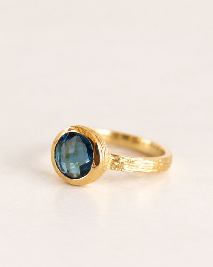 A gold ring with a textured band and a round faceted blue gemstone, photographed on a clean, light off-white background.