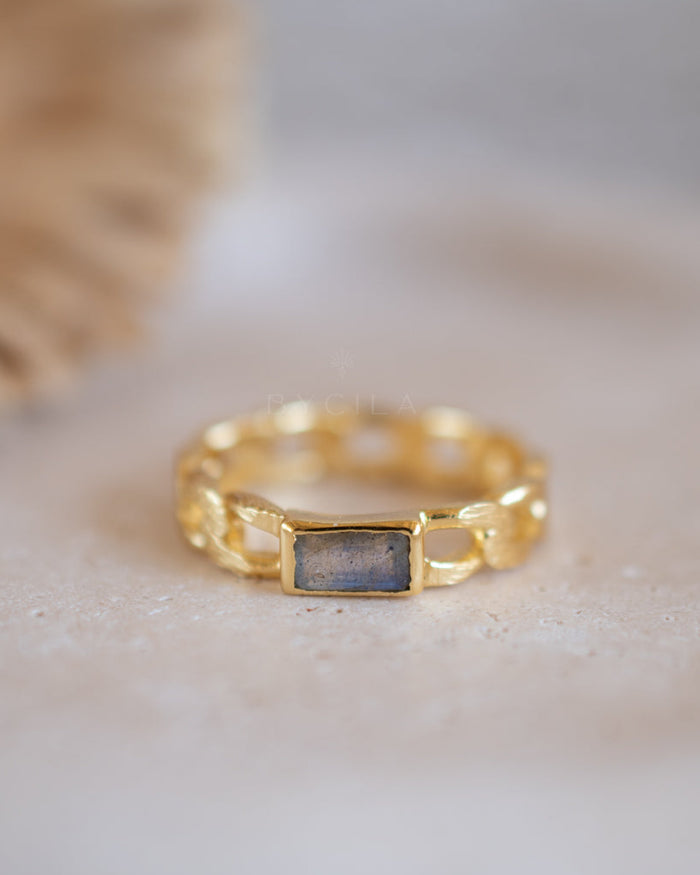 A gold chain-link ring with a rectangular gray stone in the center, resting on a textured beige surface with a soft-focus neutral background.